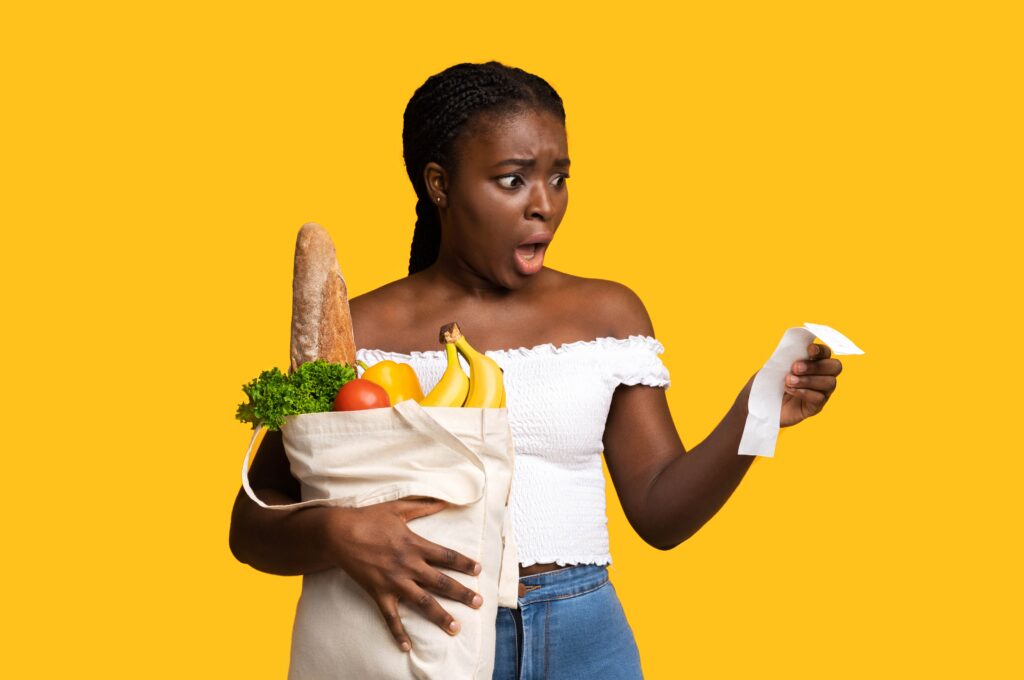 A woman with a surprised expression holding a bag of groceries and looking at a receipt. Inflation has caused the prices to increase, which is reflected in the high total amount on the receipt, affecting the value of her money.